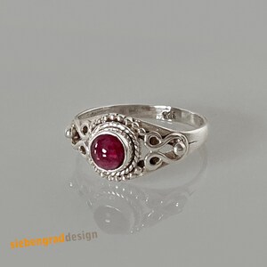 Könnte beinhalten: Ein silberner Ring mit einem verzierten Design, das einen roten Edelstein zeigt. Der Ring hat ein zartes, filigranes Muster und einen einzigen, runden, roten Stein in der Mitte.