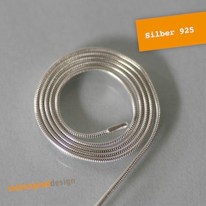 Könnte beinhalten: Eine silberne Schlangenkettenkette aus 925er Sterlingsilber. Die Kette ist auf einer grauen Oberfläche aufgerollt.