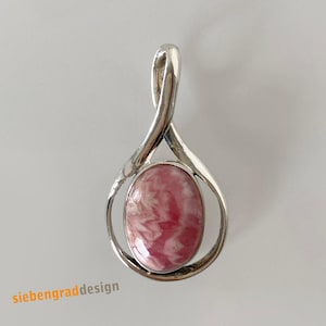 Könnte beinhalten: Ein silberner Anhänger mit einem ovalen, rosa Edelstein. Der Edelstein hat ein marmoriertes Aussehen mit Weiß- und Rosatönen. Die silberne Fassung des Anhängers hat ein einzigartiges, fließendes Design. Der Text "siebengraddesign" ist links unten sichtbar.