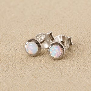 Könnte beinhalten: Ein Paar Ohrstecker aus Sterlingsilber mit schillernden, opalartigen Steinen. Die Ohrringe haben eine Lünettenfassung und werden auf sandigem Untergrund präsentiert. Der Stift eines Ohrrings ist mit "925" gestempelt.
