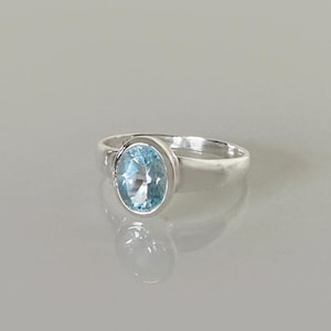 Könnte beinhalten: Ein Silberring mit einem einzelnen hellblauen Edelstein im Ovalschliff, der in einer glatten Zargenfassung sitzt. Die Ringschiene besteht aus poliertem Silber mit einem massiven, sich verjüngenden Design, das zur Mitte hin breiter wird.