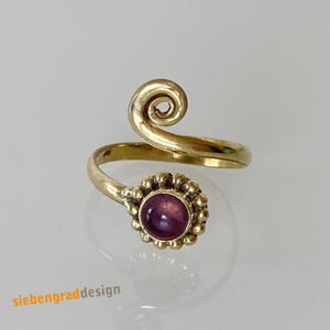 Könnte beinhalten: Ein goldener Ring mit einem violetten Edelstein und einem geschwungenen Design. Der Ring ist verstellbar und kann an jedem Finger getragen werden.