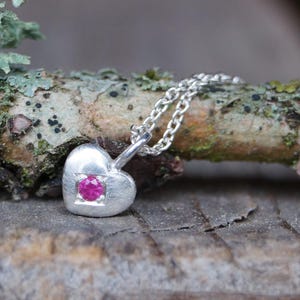 Könnte beinhalten: Eine silberne Herz-Anhänger-Halskette mit einem kleinen, quadratisch gefassten, rosa Edelstein. Der herzförmige Anhänger hängt an einer zarten Silberkette. Die Halskette wird auf einer natürlichen Holzoberfläche präsentiert.