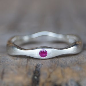 Könnte beinhalten: Ein silberner Ring mit einem wellenförmigen Band und einem kleinen, runden, rosafarbenen Rubin-Edelstein, der in der Mitte eingesetzt ist.