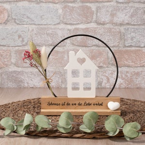 Könnte beinhalten: Weißer Keramik-Haus-Ausschnitt mit einem Herzen im Fenster, auf einem Holzsockel mit dem Text "Zuhause ist da wo die Liebe wohnt" und einem weißen Herzen. Das Haus ist von einem schwarzen Metallring umgeben.