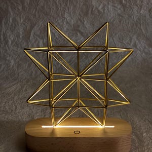 Peut inclure: Une sculpture en forme d'étoile géométrique dorée composée de fines tiges métalliques, éclairée par le bas par une bande lumineuse intégrée dans une base en bois clair de forme ovale. La base présente une icône de bouton d'alimentation.