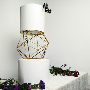 Könnte beinhalten: Ein weißer Keramiktopf mit einem goldenen geometrischen Drahtgestell oben drauf. Der Topf steht auf einem weißen Tisch mit violetten Blumen.