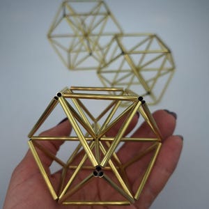 May include: Three geometric sculptures constructed from thin, hollow gold-toned metal rods. Each sculpture features a complex, multi-faceted polyhedral frame with open vertices. The structures are arranged with one in the foreground and two behind.
