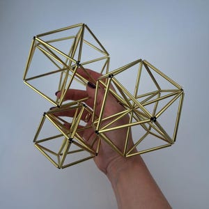 May include: Three geometric sculptures constructed from thin, metallic gold-toned rods connected at black joints. The structures feature interconnected triangular and square frames, forming complex polyhedral shapes with an open, airy design.
