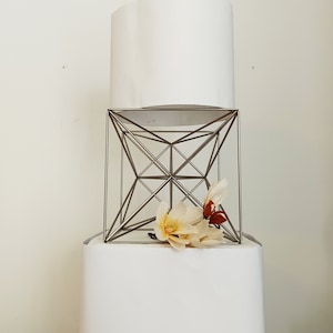 Może przedstawiać: Biały klosz lampy stoi na srebrnym, geometrycznym stojaku z białą podstawą. Na stojaku umieszczono dwie delikatne, jasnobrązowe kwiaty z czerwonymi akcentami.