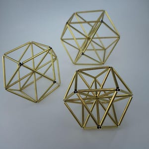 May include: Three geometric sculptures constructed from thin, metallic gold-colored rods. Each object features a complex, multi-faceted polyhedral frame with open spaces between the connected bars, creating a three-dimensional wireframe structure.