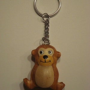 Könnte beinhalten: Ein Holz-Affen-Schlüsselanhänger mit braunem Körper, beigefarbenem Bauch und schwarzen Augen. Der Affe ist an einem silbernen Schlüsselring und einer Kette befestigt.