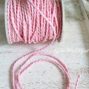 Könnte beinhalten: Eine Spule aus rosa geflochtenem Kordel. Die Kordel ist fest auf einer weißen Kunststoffspule aufgewickelt. Ein Stück der Kordel ist abgewickelt und liegt vor der Spule. Die Kordel hat eine weiche, pastellrosa Farbe. Das Bild ist gut beleuchtet und zeigt die Textur der Kordel.