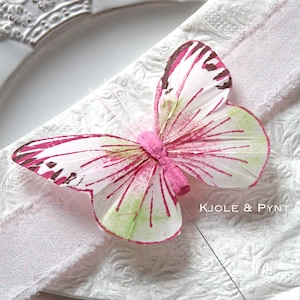 Könnte beinhalten: Ein dekorativer Schmetterling mit weißen Flügeln, akzentuiert mit rosa, braunen und grünen Details, liegt auf einer weißen Serviette. Der Schmetterling ist auf einem weißen Teller zentriert, mit einem rosa Band und dem Text "KJOLE & PYNT".