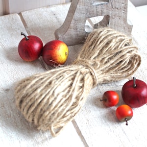 Könnte beinhalten: Ein rustikales Stillleben mit einem Garnknäuel, mehreren roten und gelben Äpfeln und einem Holzbuchstaben "A". Das Garn ist mit einer Schnur aus Naturfaser gebunden. Die Äpfel haben dunkle Stiele. Der Text "KJOLE & PYNT" ist sichtbar.