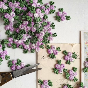 Könnte beinhalten: Nahaufnahme von gestickten Blumenapplikationen in Lavendel, Weiß und Grün. Die Applikationen sind auf einer weißen Oberfläche verstreut, mit einer Schere und einem Vintage-Papier mit dem Wort "Paris" sichtbar.