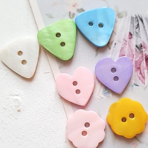 Könnte beinhalten: Eine Sammlung bunter Knöpfe in verschiedenen Formen, darunter Dreiecke, Herzen und eine Blütenform. Die Knöpfe sind in Weiß-, Grün-, Blau-, Rosa-, Lila- und Gelbtönen gehalten. Jeder Knopf hat zwei Löcher zum Nähen.