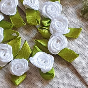 Könnte beinhalten: Nahaufnahme von handgefertigten weißen Satinbandrosen mit grünen Satinbandblättern. Die floralen Verzierungen sind auf einem strukturierten, neutralfarbenen Stoff angeordnet. Eine kleine Schere befindet sich im Vordergrund.
