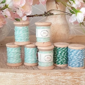 Könnte beinhalten: Holzspulen mit Faden in verschiedenen Farben, darunter Türkis, Grün und Blau, auf einem Holztablett angeordnet. Einige Spulen tragen die Aufschrift "5m". Im Hintergrund eine Vase mit rosa Blüten.