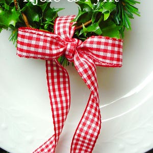 Può includere: Una ghirlanda festiva con foglie verdi e un fiocco a scacchi rosso e bianco. La ghirlanda presenta il testo "Kjole & Pynt" in bianco. Una bacca rossa è in cima. La ghirlanda è su un piatto bianco.