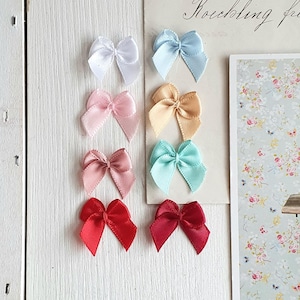 Könnte beinhalten: Eine Sammlung von acht kleinen Satin-Schleifen in verschiedenen Farben, darunter Weiß, Hellblau, Rosa, Pfirsich, Türkis und Rot. Die Schleifen sind auf einer weißen Holzoberfläche angeordnet, mit einer Vintage-Postkarte und einem Blumendruck im Hintergrund.