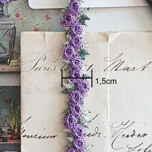Könnte beinhalten: Ein dekorativer Besatz mit einer Reihe von lila rosenartigen Verzierungen, jede etwa 1,5 cm im Durchmesser, verbunden durch grüne Blätter. Der Besatz wird vor einem Vintage-Papierhintergrund präsentiert.