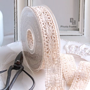 Könnte beinhalten: Eine Rolle cremefarbenes Spitzenband mit einem filigranen Blumenmuster. Das Band ist teilweise abgerollt und zeigt sein aufwendiges Design. Eine antike schwarze Schere liegt neben dem Band auf einem weißen Teller, mit einem weißen Fotorahmen im Hintergrund.