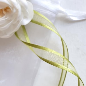 Könnte beinhalten: Nahaufnahme eines weißen Blumenarrangements mit Perlenakzent und einem grün-goldenen Band. Das Band ist gewellt und verleiht dem Arrangement eine dekorative Note. Der Hintergrund ist ein weicher weißer Stoff.