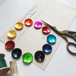 Könnte beinhalten: Eine Sammlung schillernder, farbenfroher Knöpfe, die auf einem Vintage-Papier kreisförmig angeordnet sind. Die Knöpfe sind in Gelb-, Orange-, Rosa-, Rot-, Türkis-, Blau-, Grün- und Schwarztönen gehalten. Eine goldene Schere und Garnrollen sind ebenfalls vorhanden.