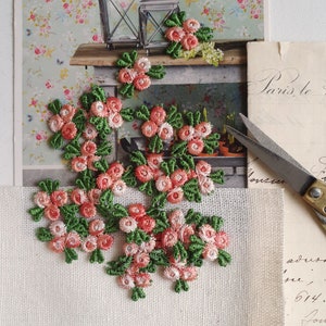 Könnte beinhalten: Gestickte Blumenapplikationen mit Büscheln aus rosa und pfirsichfarbenen Rosen mit grünen Blättern. Die Applikationen sind auf einer weißen Stoffoberfläche angeordnet, mit einer Vintage-Postkarte und einer Schere im Hintergrund. Ideal für Bastelarbeiten und Verzierungen.