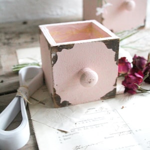 Könnte beinhalten: Zwei kleine, rosafarbene Holzschubladen mit runden Knöpfen. Die Boxen haben eine rustikale, Vintage-Ästhetik mit sichtbarer Holzmaserung und gealterten Kanten. Eine Box ist im Fokus, mit einem Bündel weißem Papier und getrockneten Blumen in der Nähe.