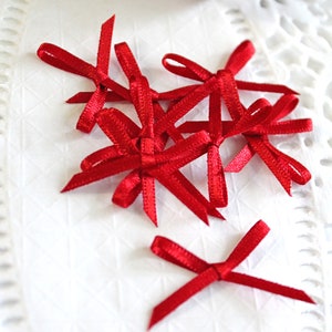Könnte beinhalten: Kleine, leuchtend rote Satin-Schleifen auf einer weißen, strukturierten Oberfläche. Im Hintergrund ist eine Rolle rot-weißes Garn zu sehen. Die zarten Schleifen eignen sich ideal zum Basteln oder zum Verpacken von Geschenken.