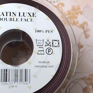 Puede incluir: Un carrete de cinta Satin Luxe Double Face marr&oacute;n oscuro. La etiqueta indica que es 100% PES y est&aacute; fabricado en Suiza. Tambi&eacute;n se ven las instrucciones de lavado. La marca "Kjole & Pynt" est&aacute; en la parte inferior.