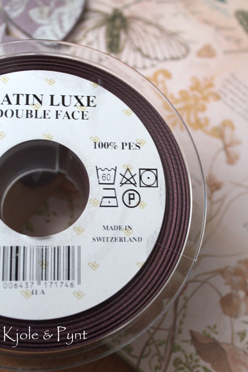 Puede incluir: Un carrete de cinta Satin Luxe Double Face marr&oacute;n oscuro. La etiqueta indica que es 100% PES y est&aacute; fabricado en Suiza. Tambi&eacute;n se ven las instrucciones de lavado. La marca "Kjole & Pynt" est&aacute; en la parte inferior.