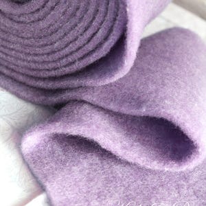 Könnte beinhalten: Nahaufnahme einer Rolle weichen, lavendelfarbenen Filzstoffs. Der Filz ist dick und hat eine strukturierte Oberfläche. Der Stoff ist aufgerollt, wobei das Ende der Rolle sichtbar ist. Das Bild zeigt die Textur und Farbe des Filzes.