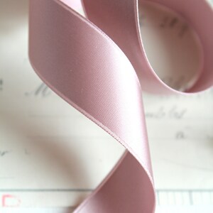 Puede incluir: Primer plano de una cinta de raso rosa p&aacute;lido, elegantemente curvada. La cinta tiene una textura suave y brillante, sobre un fondo borroso blanquecino. Ideal para manualidades, envolver regalos o fines decorativos.