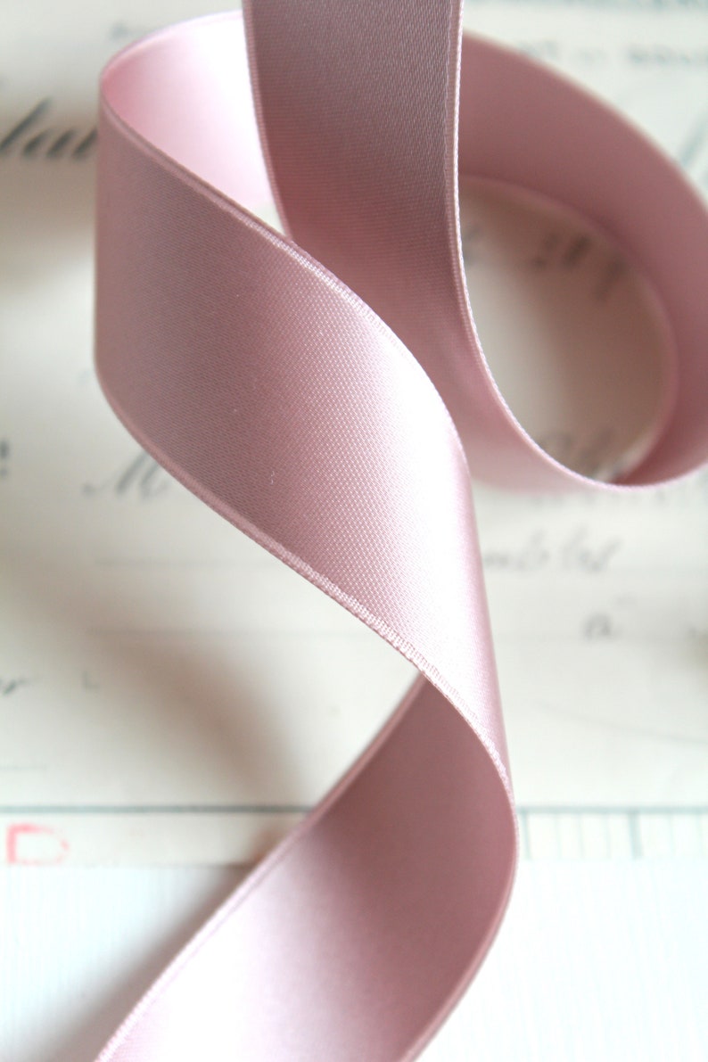 Puede incluir: Primer plano de una cinta de raso rosa p&aacute;lido, elegantemente curvada. La cinta tiene una textura suave y brillante, sobre un fondo borroso blanquecino. Ideal para manualidades, envolver regalos o fines decorativos.