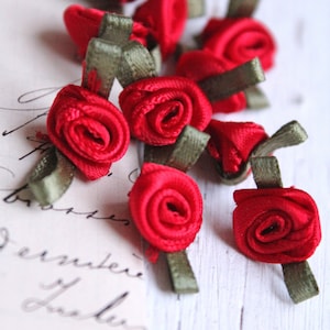 Könnte beinhalten: Nahaufnahme von mehreren kleinen, roten Satinrosen mit olivgrünen Bandstielen. Die Rosen sind eng gewickelt, und das Band hat eine flache, rechteckige Form. Der Hintergrund ist eine verschwommene, cremefarbene Oberfläche.