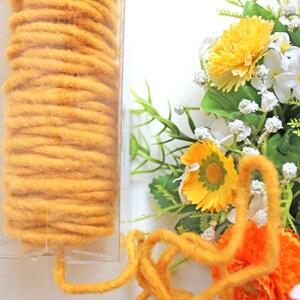 May include: A clear plastic container holds a tightly wound roll of mustard-yellow yarn. A loose strand of the same yarn is in the foreground, with a bouquet of yellow and orange flowers in the background. The text "Kjole & Pynt" is visible.