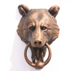 Puede incluir: Aldaba de puerta con forma de cabeza de oso en color bronce con un asa en forma de anillo. La cara del oso está detallada con textura de pelaje y rasgos faciales. La aldaba está montada sobre una superficie blanca.