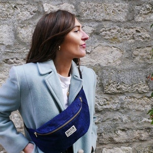 May include: A woman wearing a light blue coat with a dark blue denim fanny pack. The fanny pack has a zipper and the text "MAISON BOISGU" printed on it.