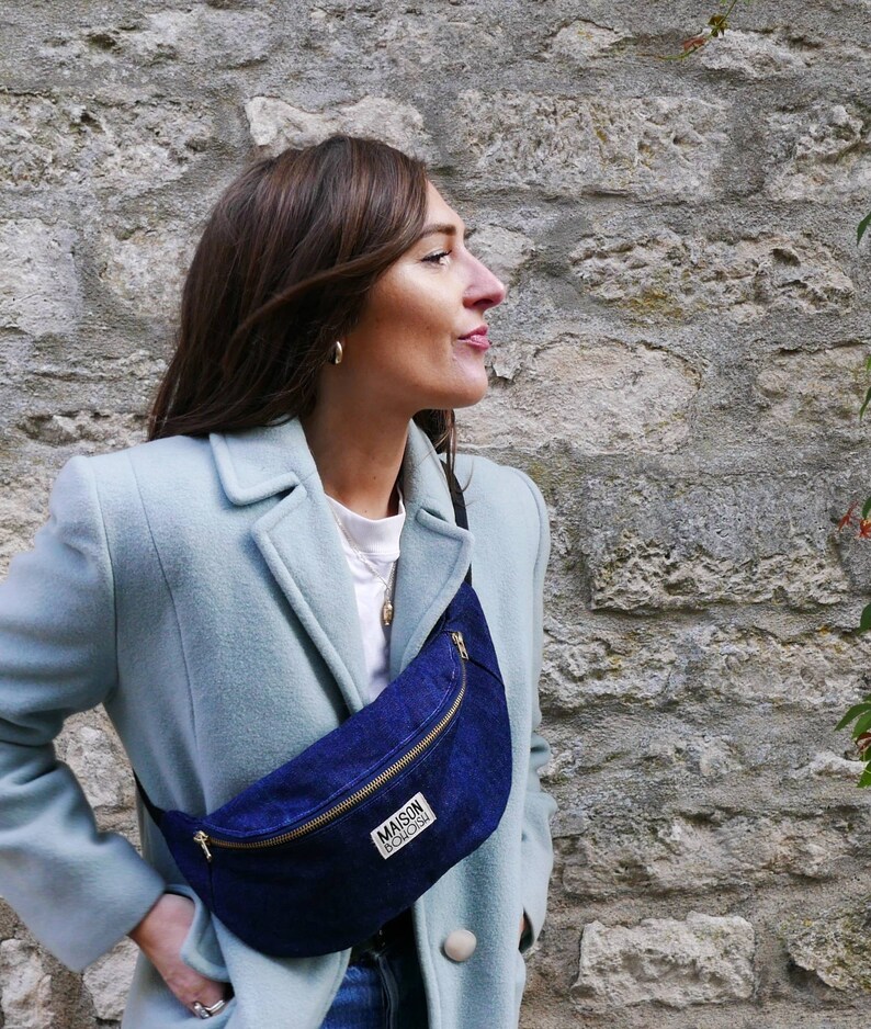 May include: A woman wearing a light blue coat with a dark blue denim fanny pack. The fanny pack has a zipper and the text "MAISON BOISGU" printed on it.
