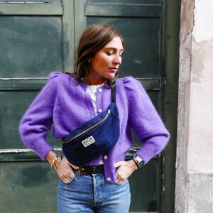 May include: A woman wearing a purple fuzzy cardigan, blue jeans, and a black fanny pack with the text "Maison Scotch" on it.
