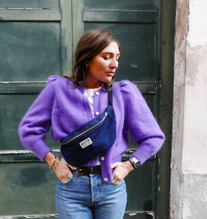 May include: A woman wearing a purple fuzzy cardigan, blue jeans, and a black fanny pack with the text "Maison Scotch" on it.
