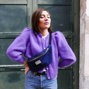 May include: A woman wearing a purple fuzzy cardigan with a blue denim fanny pack that says "Maison Standards" in white lettering. She is wearing blue jeans and a white t-shirt.