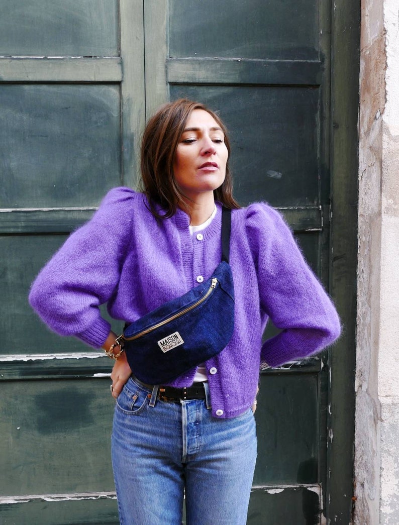 May include: A woman wearing a purple fuzzy cardigan with a blue denim fanny pack that says "Maison Standards" in white lettering. She is wearing blue jeans and a white t-shirt.