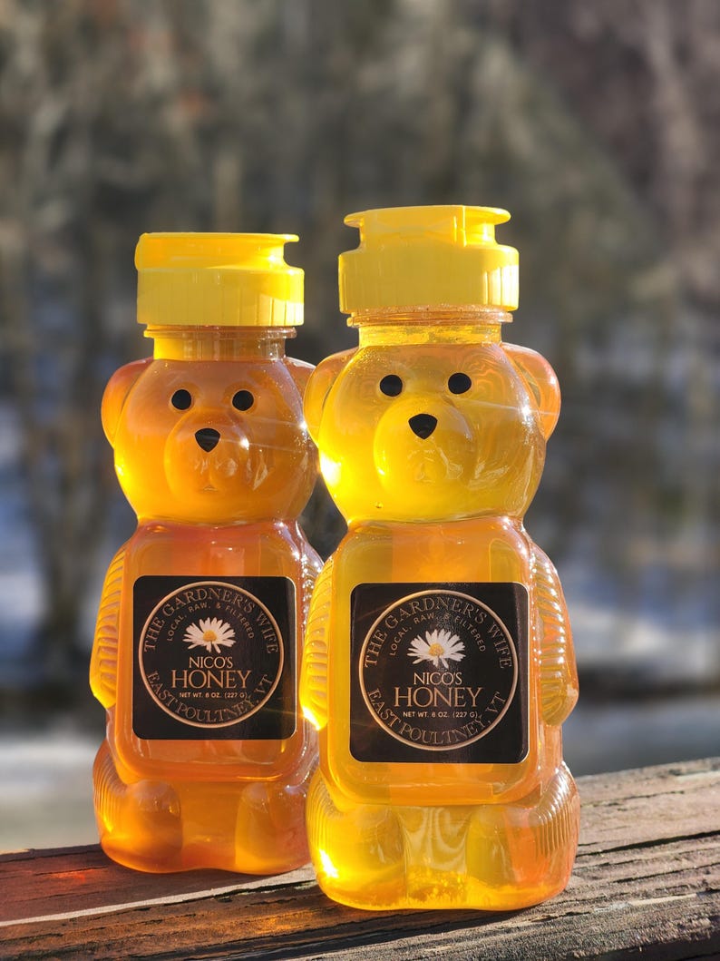 May include: Two bear-shaped honey bottles filled with golden honey. The bottles have yellow caps and black labels with white text that reads "The Gardner's Wife Nico's Honey". Each bottle contains 8 oz (227 g) of honey.