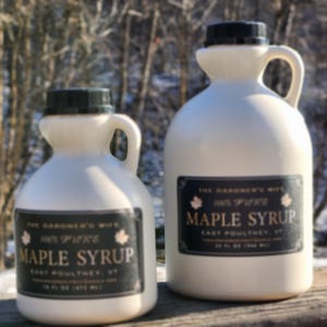 Puede incluir: Dos jarras de plástico blanco de jarabe de arce con tapas y asas negras. Cada jarra tiene una etiqueta negra con texto blanco que dice "The Gardener's Wife 100% Pure Maple Syrup East Poultney, VT". La jarra más grande contiene 946 ml.