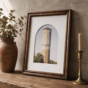 May include: A framed print of a water tower with a brown wooden frame. The print is set against a white mat. A brown vase with greenery and a brass candlestick with a lit candle are on a wooden surface.