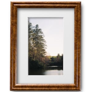 May include: A framed photograph of a lake surrounded by trees. The lake is calm and reflects the sky. The trees are green and brown. The frame is brown wood with a white mat.